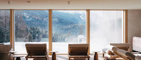 Amplia habitación con grandes ventanales que ofrecen vistas a la montaña, suelo de madera y muebles modernos, incluyendo un sofá y sillones.