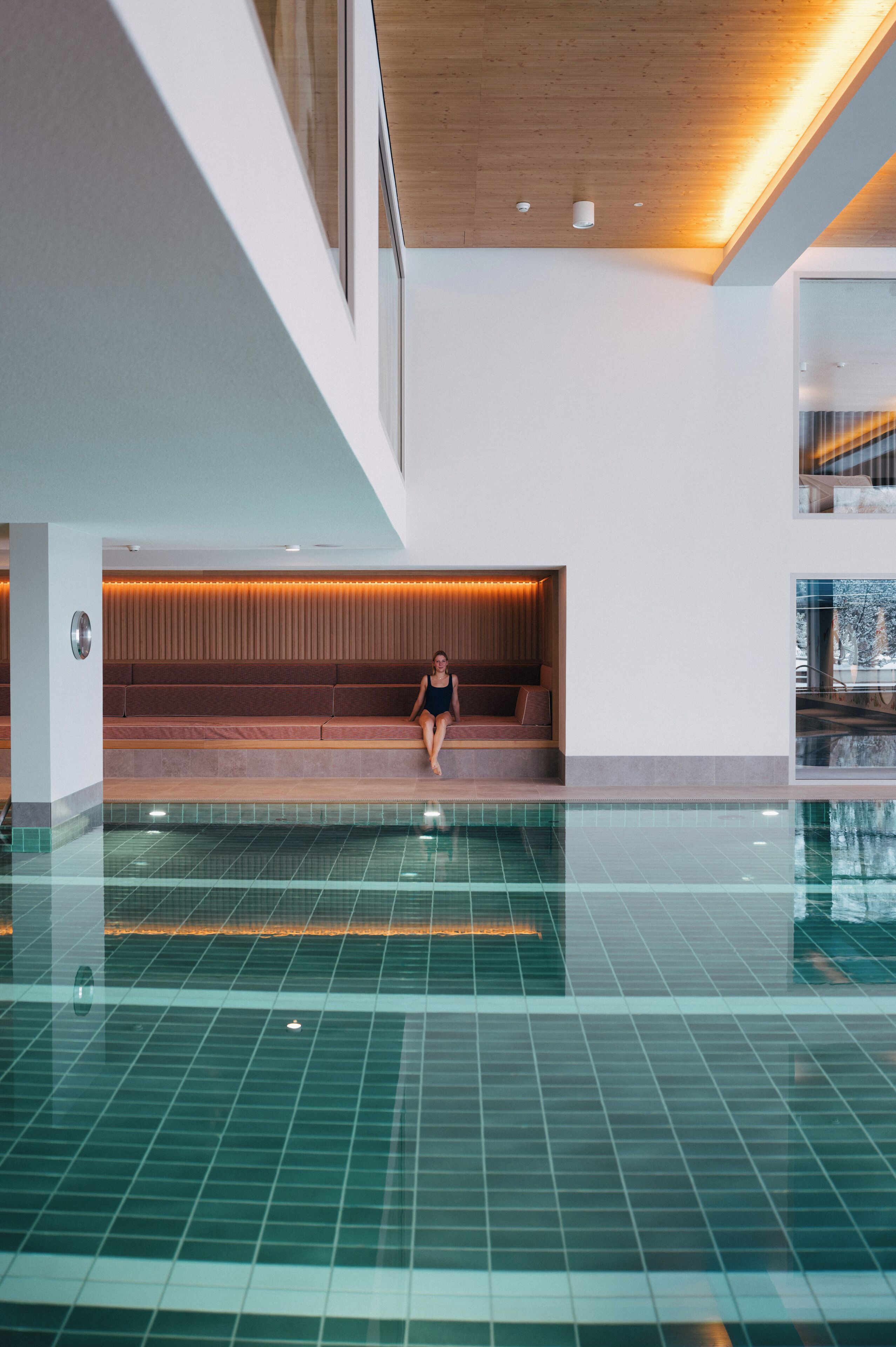 A modern indoor swimming pool with a wooden ceiling and a seating area with a view of the outside.