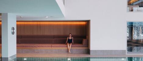 A modern indoor swimming pool with a wooden ceiling and a seating area with a view of the outside.
