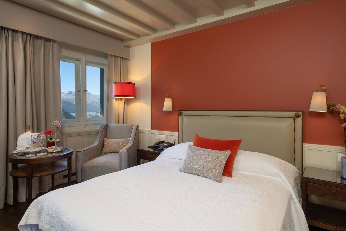 A hotel room with a bed, a chair, a table with a bottle and glasses, and a lamp.