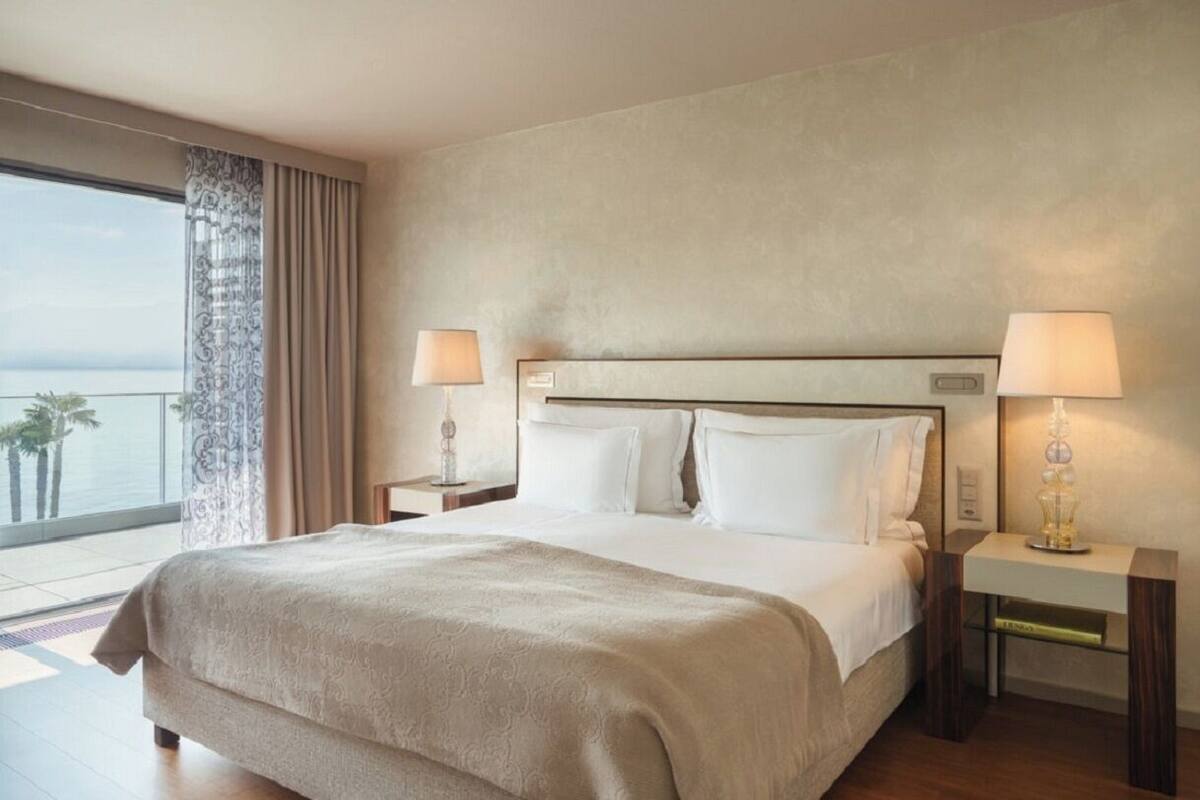 A hotel room with a large bed, two bedside tables with lamps, a view of the sea, and a balcony with curtains.