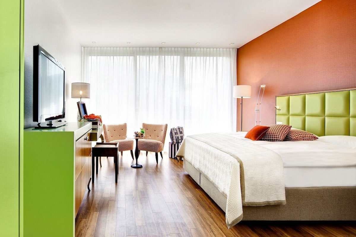 A hotel room with a bed, a desk with a computer, two armchairs, a TV, and a window with curtains.