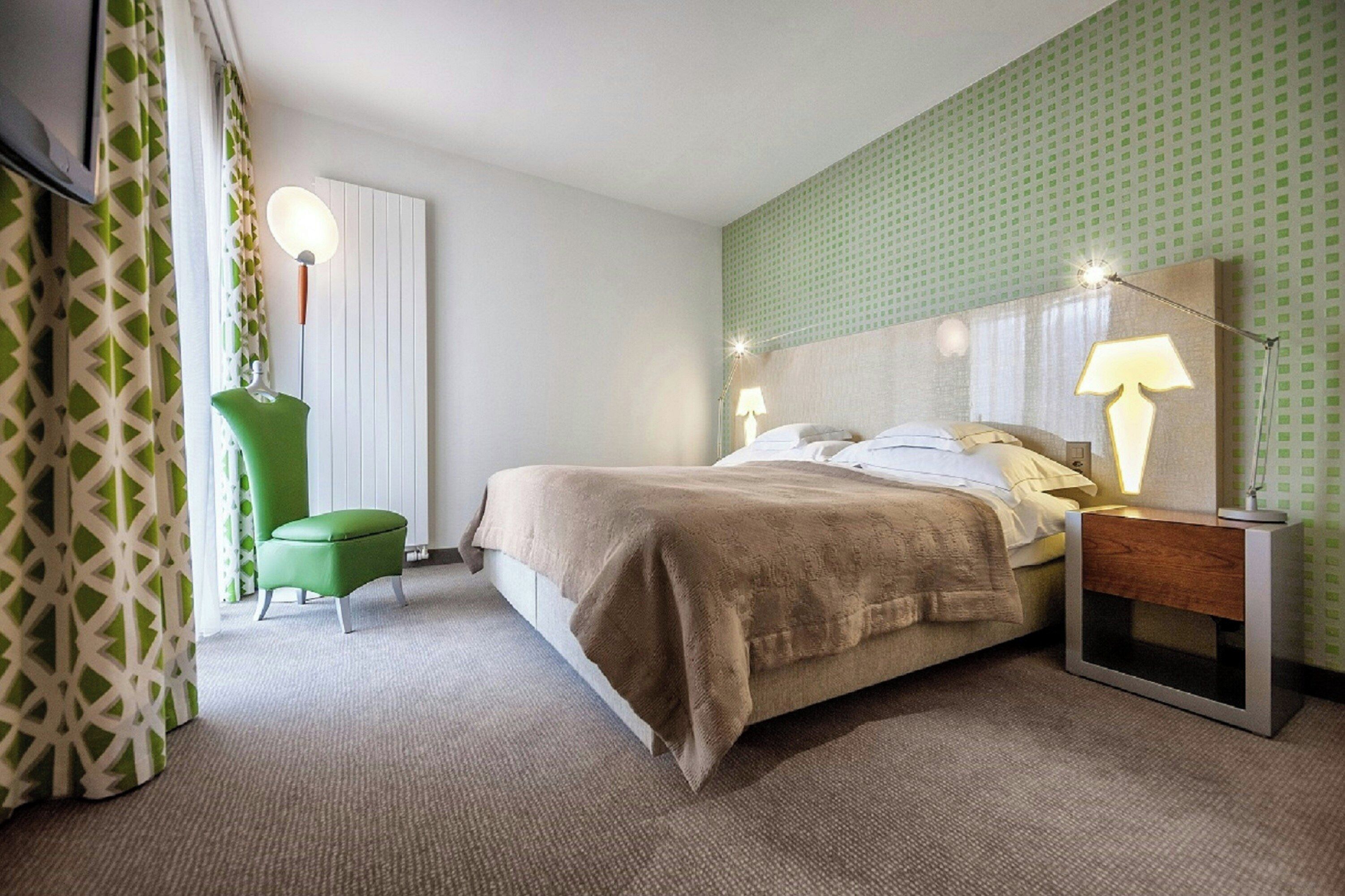 A hotel room with a bed, bedside lamps, a green chair, and a patterned curtain.