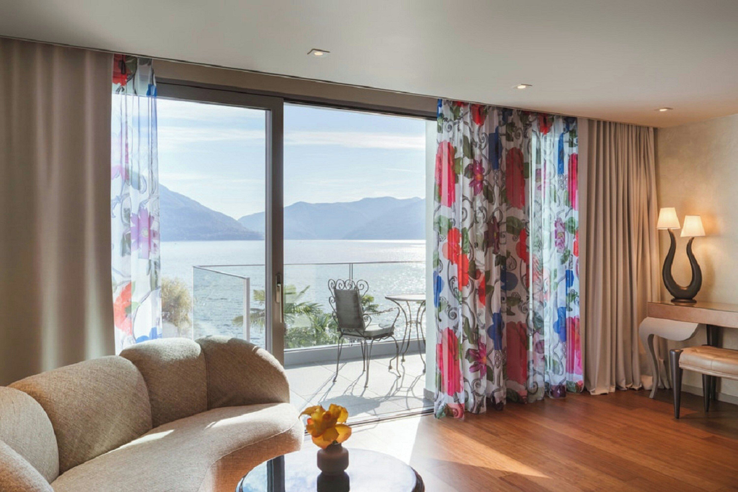 Une pièce comprenant un canapé, une table avec un vase, un balcon avec une table et des chaises, et une vue sur un lac et des montagnes.