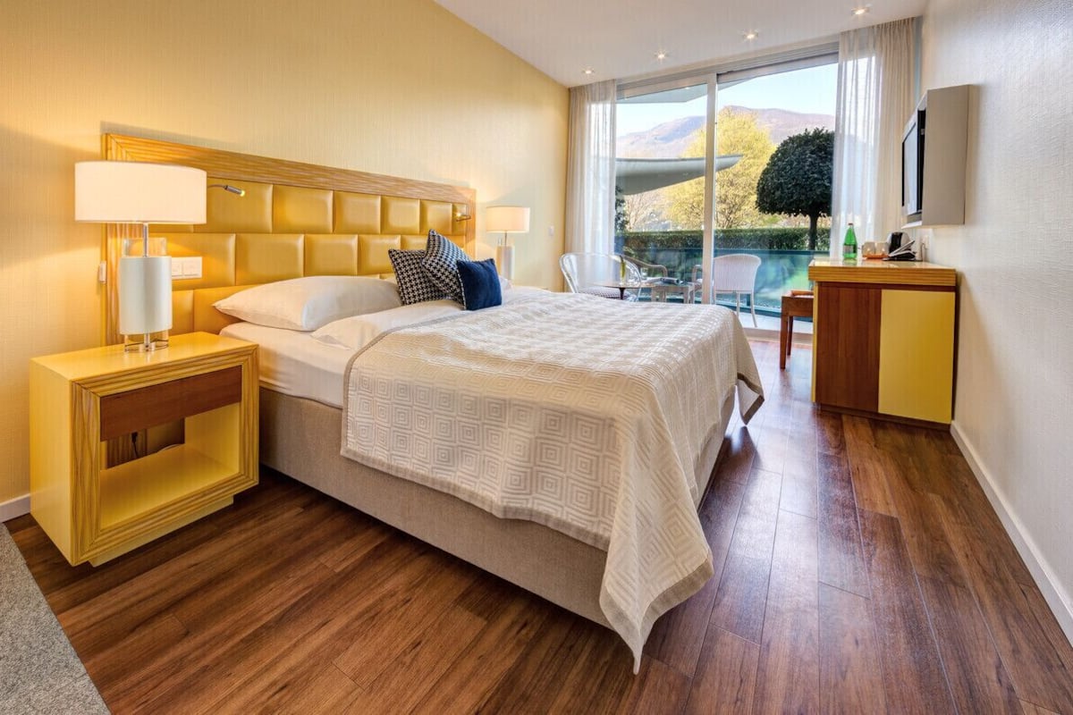 A hotel room with a large bed, a nightstand, a desk, a chair, and a view of the outdoors.