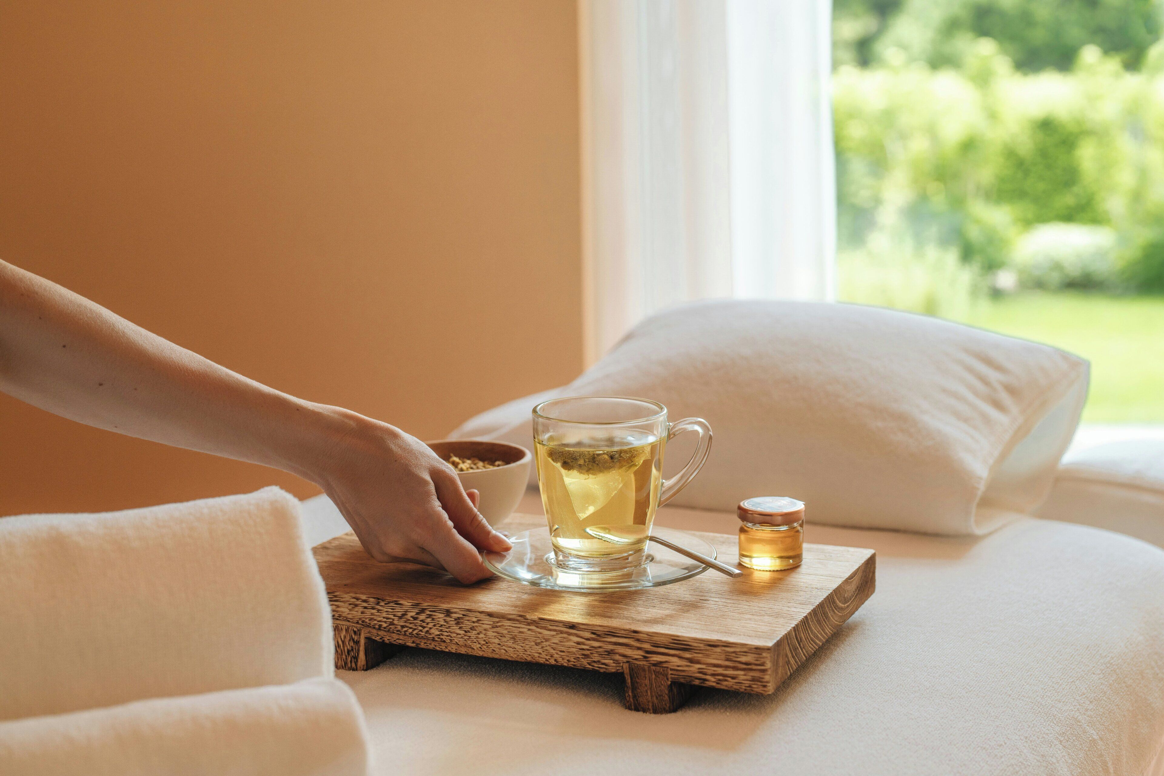 A hand placing a glass mug on a wooden tray on a bed with pillows.