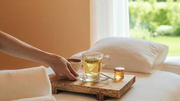 A hand placing a glass mug on a wooden tray on a bed with pillows.