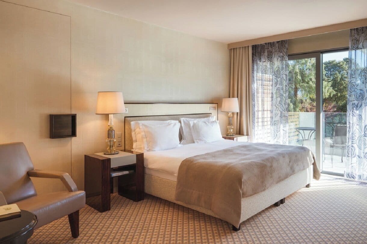 Habitación de hotel con una cama grande, dos mesitas de noche con lámparas, una silla y vistas al exterior a través de una puerta corrediza de vidrio.