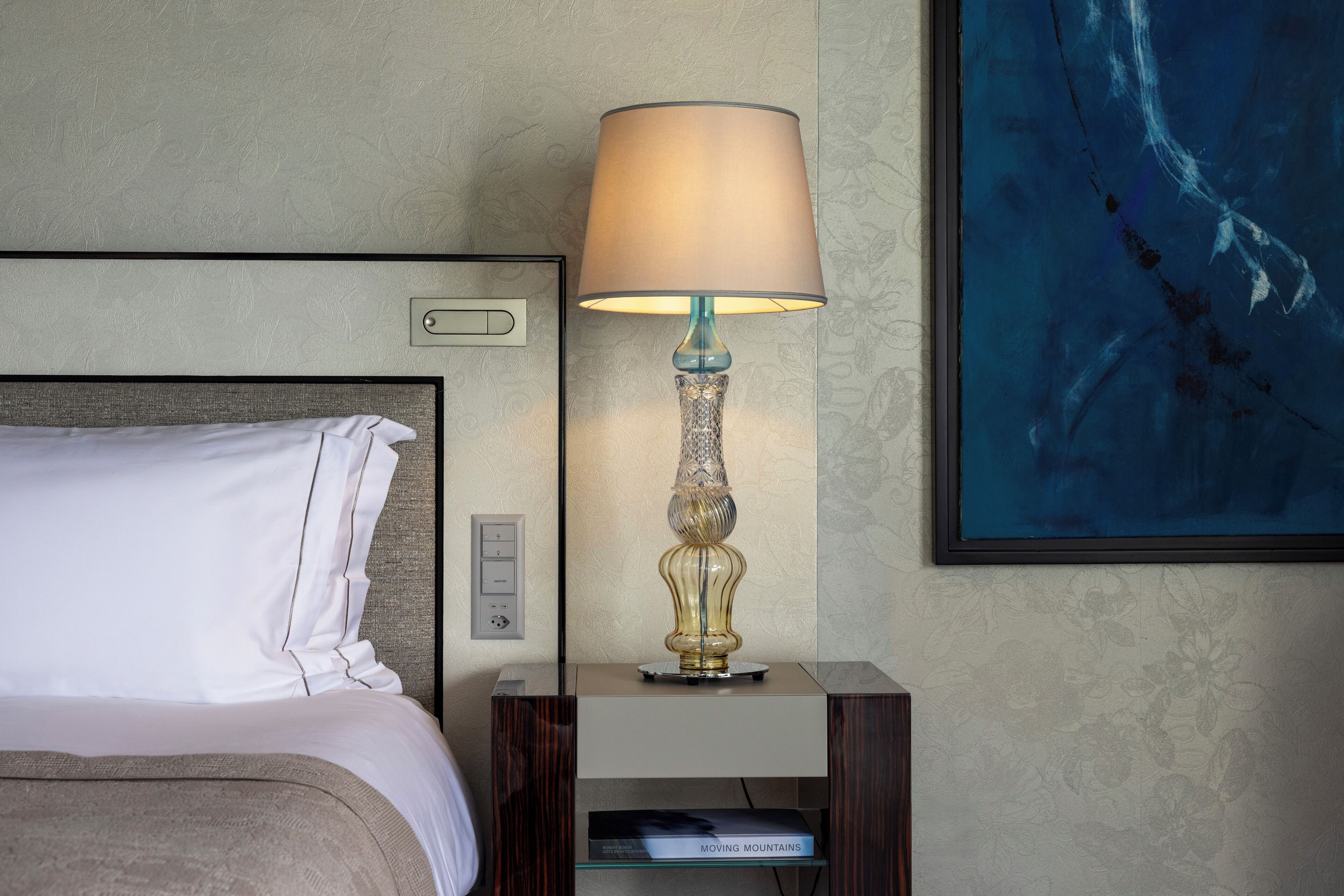 A neatly made bed with white linens, a decorative lamp on a bedside table, and a framed artwork on the wall.