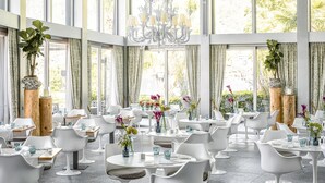 A spacious dining area with white tables and chairs, large windows, and potted plants. - Hotel Eden Roc (Ascona)