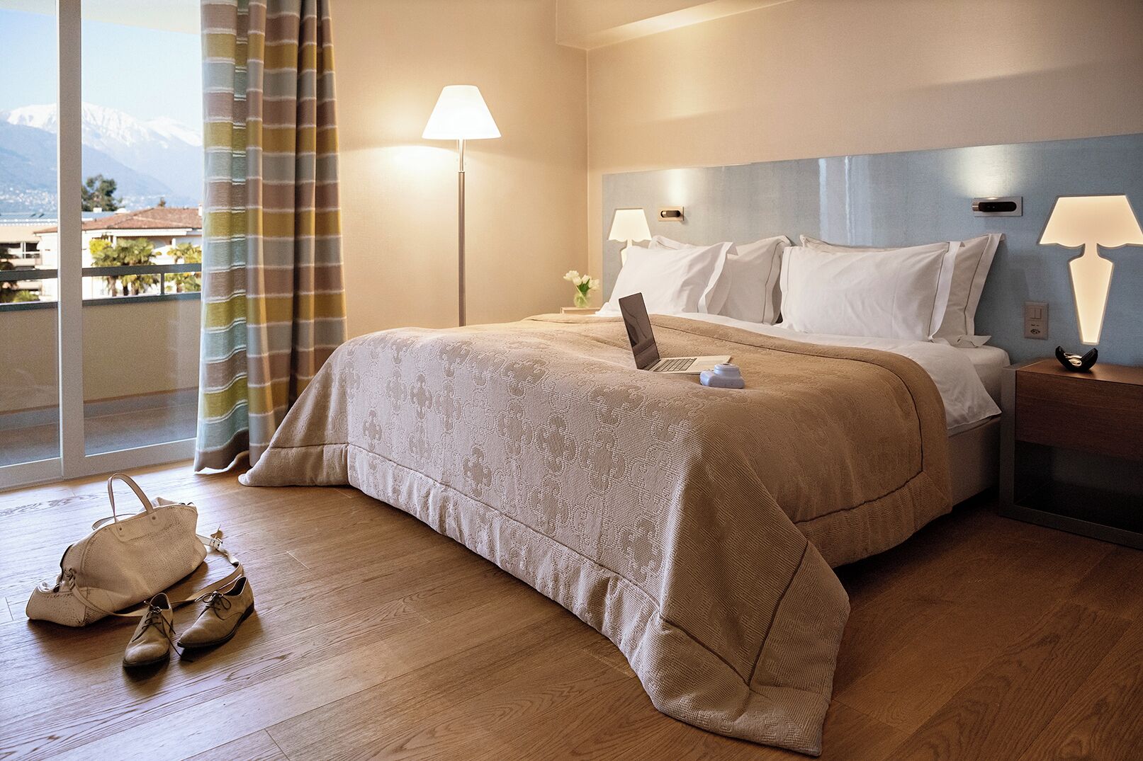 Hotelkamer met een bed, een laptop op het bed, een nachtkastje met een lamp, een paar schoenen op de vloer, en een uitzicht op bergen door het raam.