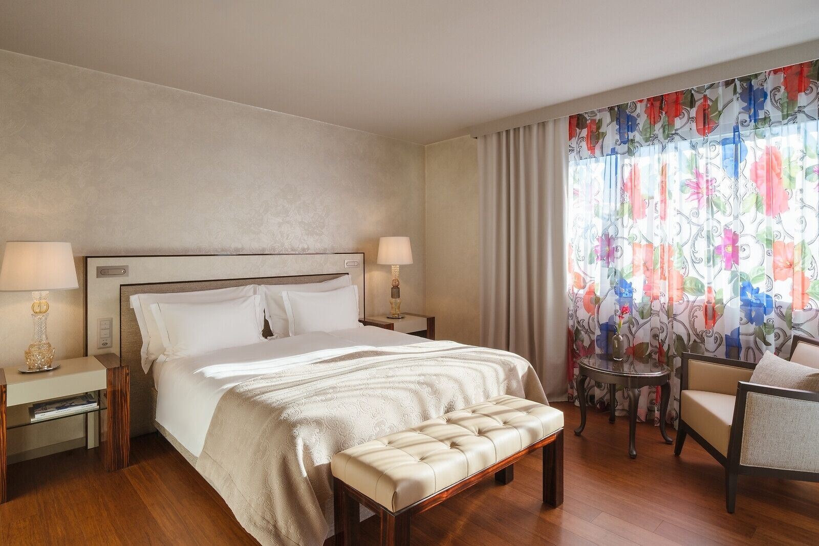 Une chambre d’hôtel avec un grand lit, deux tables de chevet avec des lampes, une petite table ronde, une chaise et une fenêtre agrémentée de rideaux à motifs floraux.