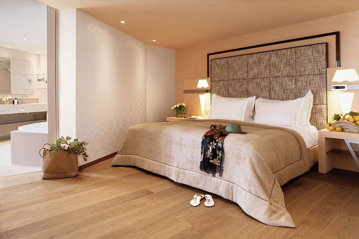 A modern hotel room with a large bed, a bedside table with a lamp, a small bedside table with a lamp, a large window, and a bathroom visible in the background.