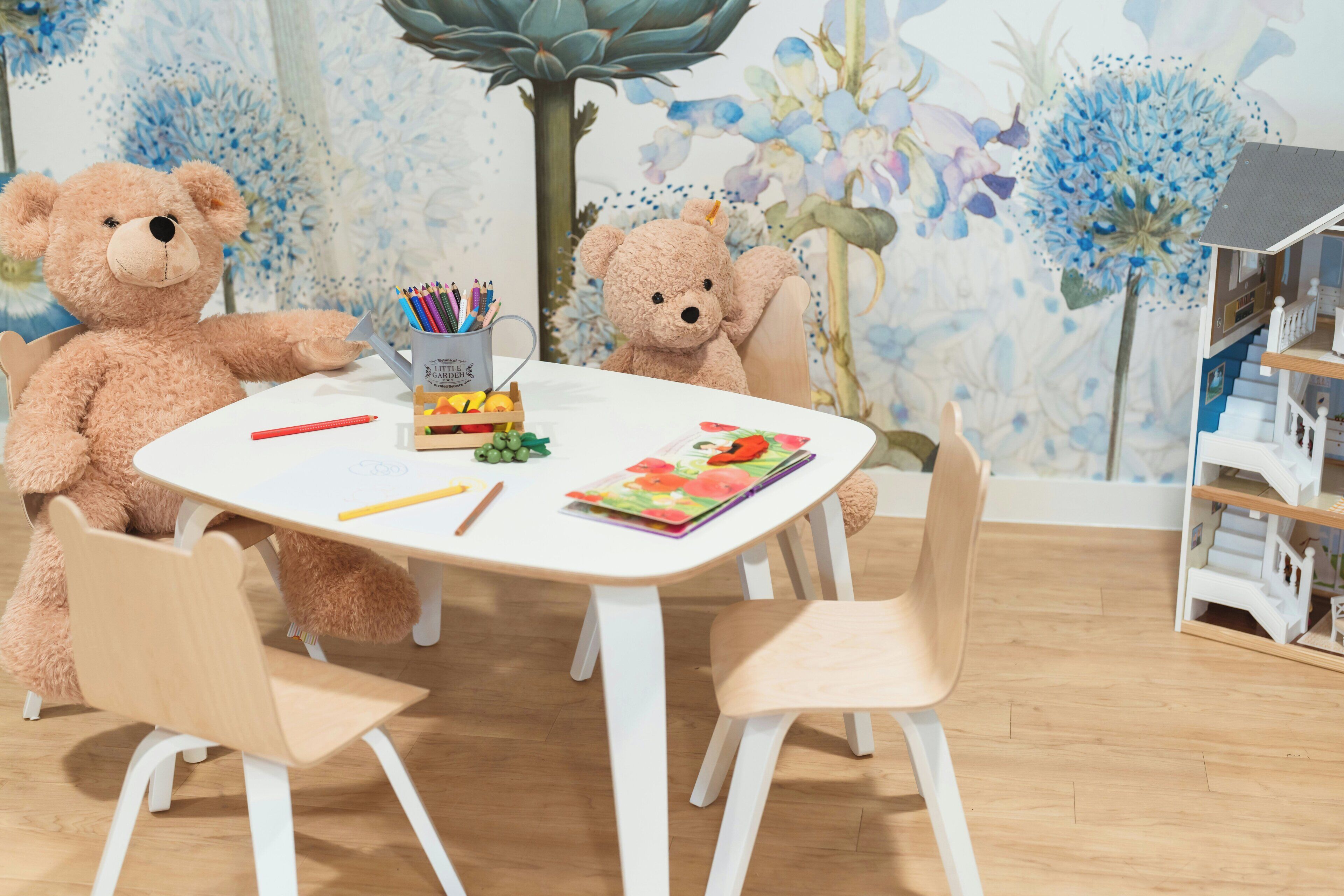 A room with a white table, two chairs, a teddy bear, a cup with colored pencils, and a book.