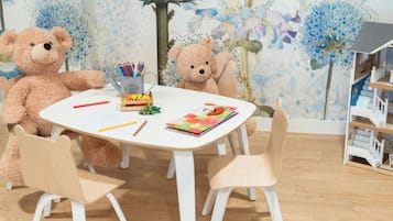 A room with a white table, two chairs, a teddy bear, a cup with colored pencils, and a book.