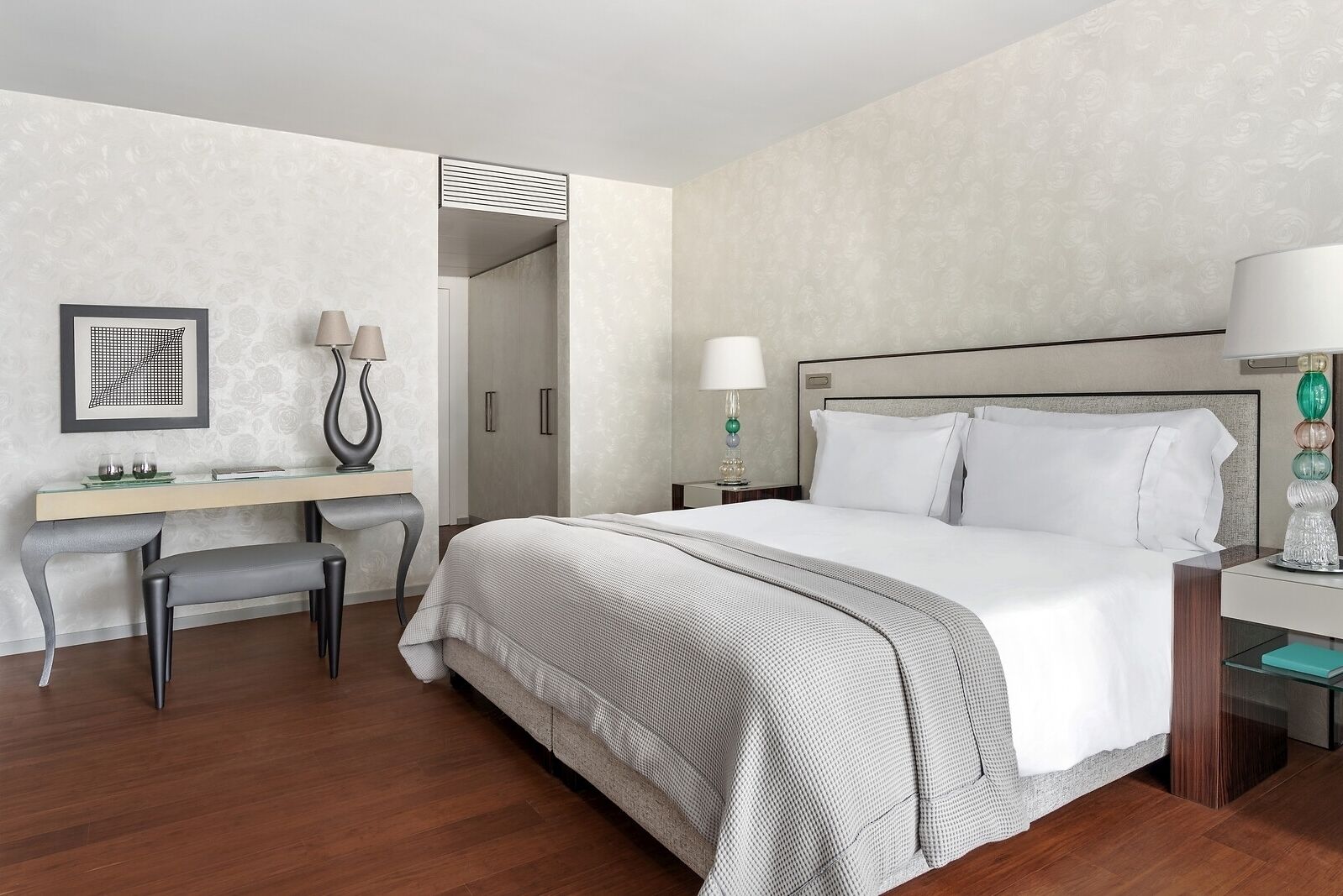 A hotel room with a large bed, a desk with a chair, a nightstand, and a wall-mounted lamp.