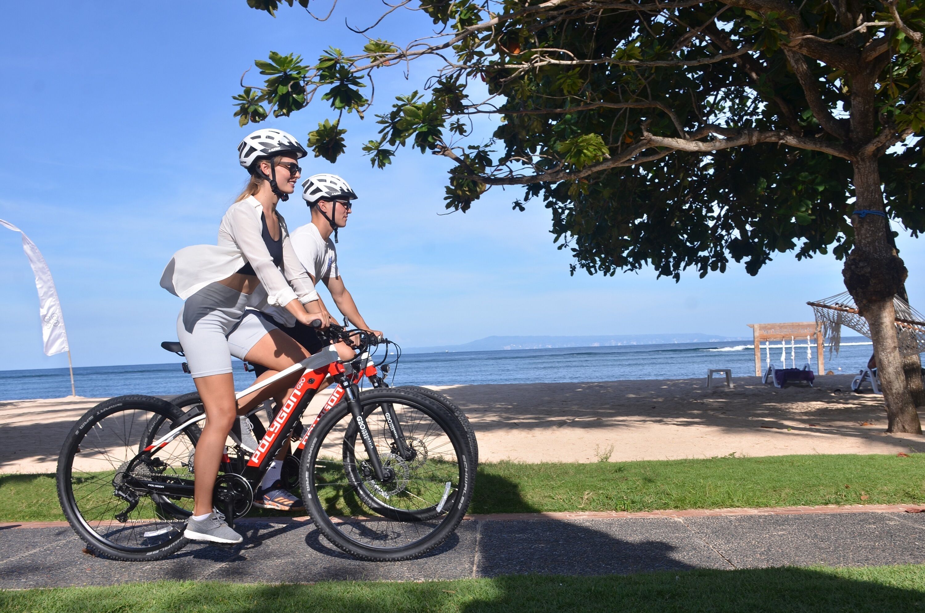 Deux personnes à vélo, près d’une plage, avec un arbre et une structure en bois.