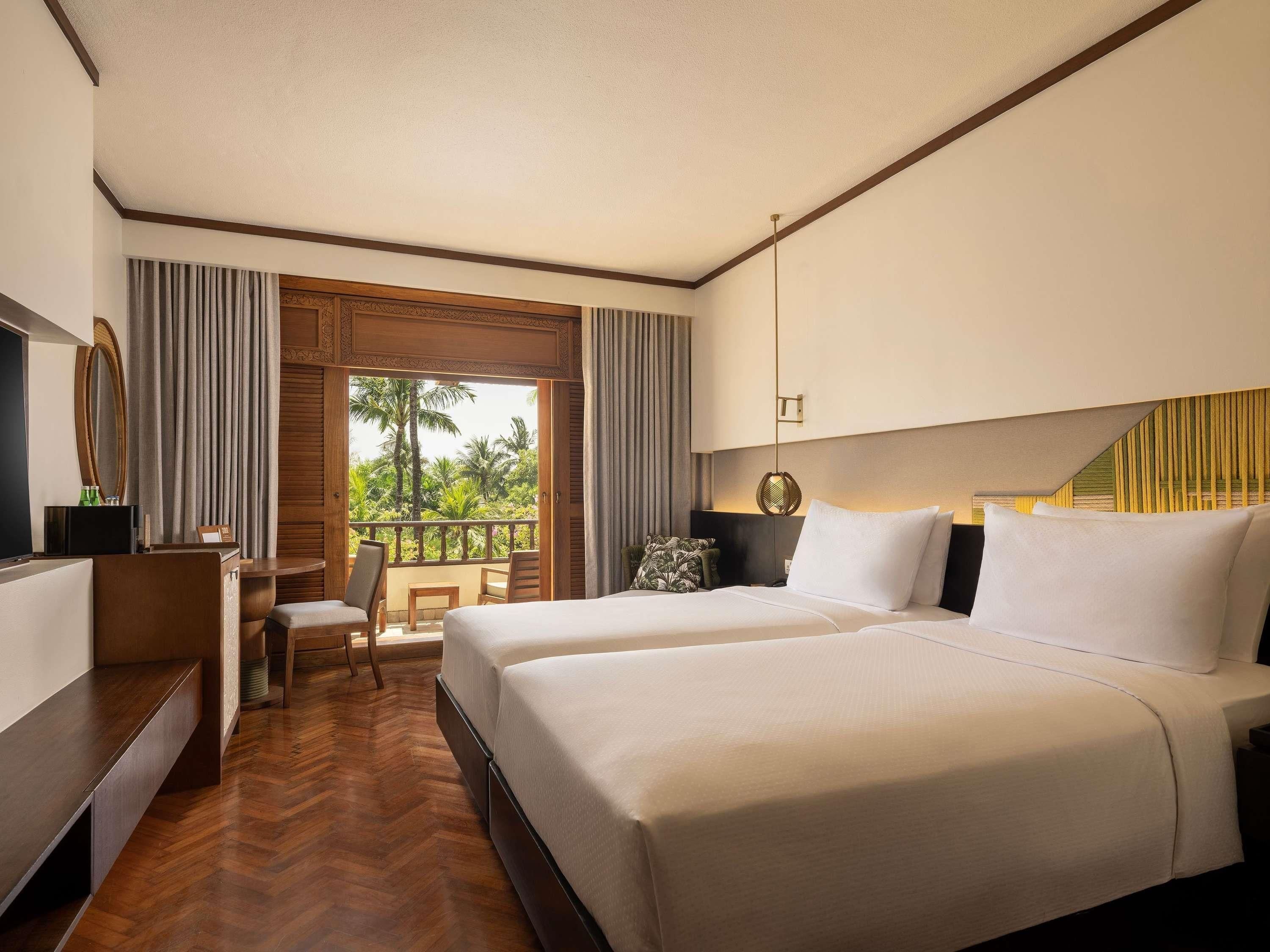 A hotel room with two beds, a desk, a chair, and a balcony with a view of palm trees.