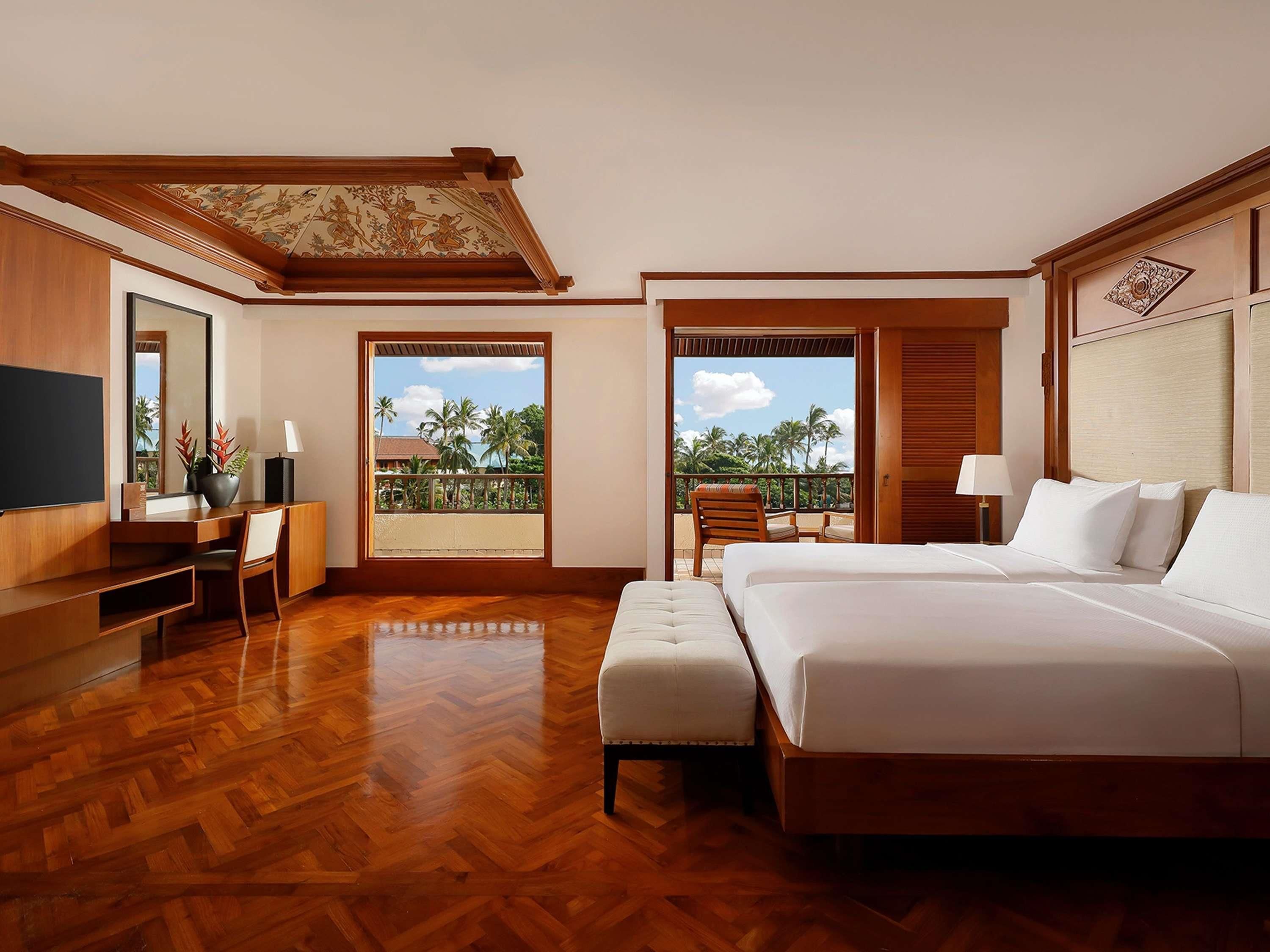A hotel room with a large bed, a desk, a chair, and a balcony with a view of palm trees.