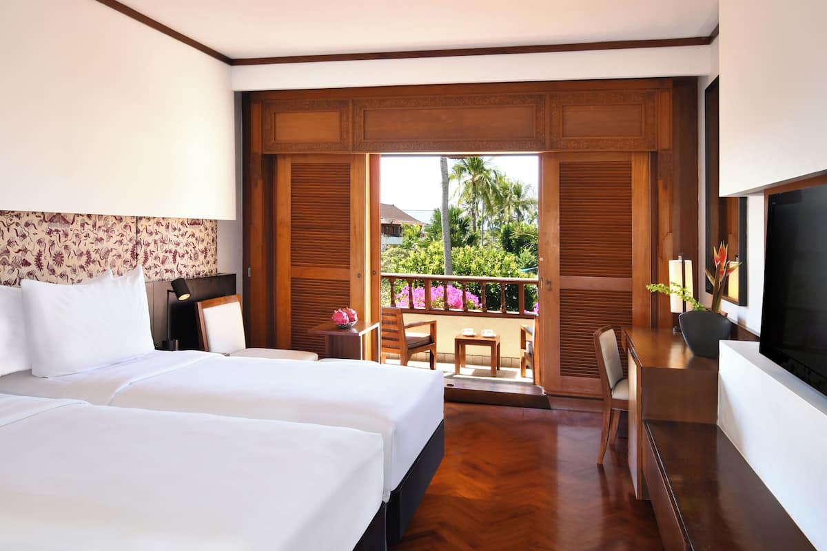 A hotel room with two beds, a desk, a TV, and a balcony with a view of palm trees and flowers.