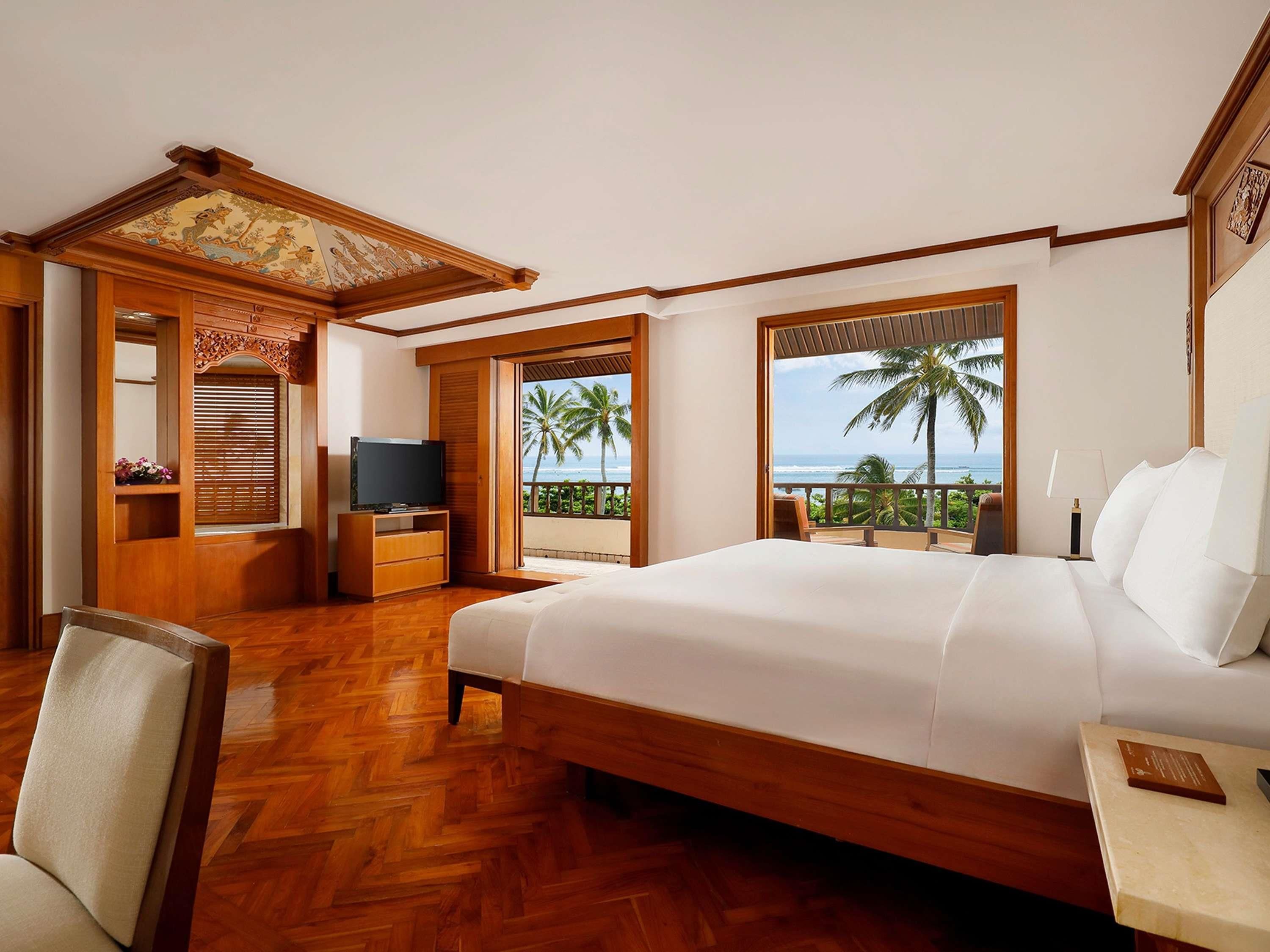 Ein Hotelzimmer mit einem großen Bett, Holzboden, einem Fernseher und einem Balkon mit Blick auf Palmen und das Meer.