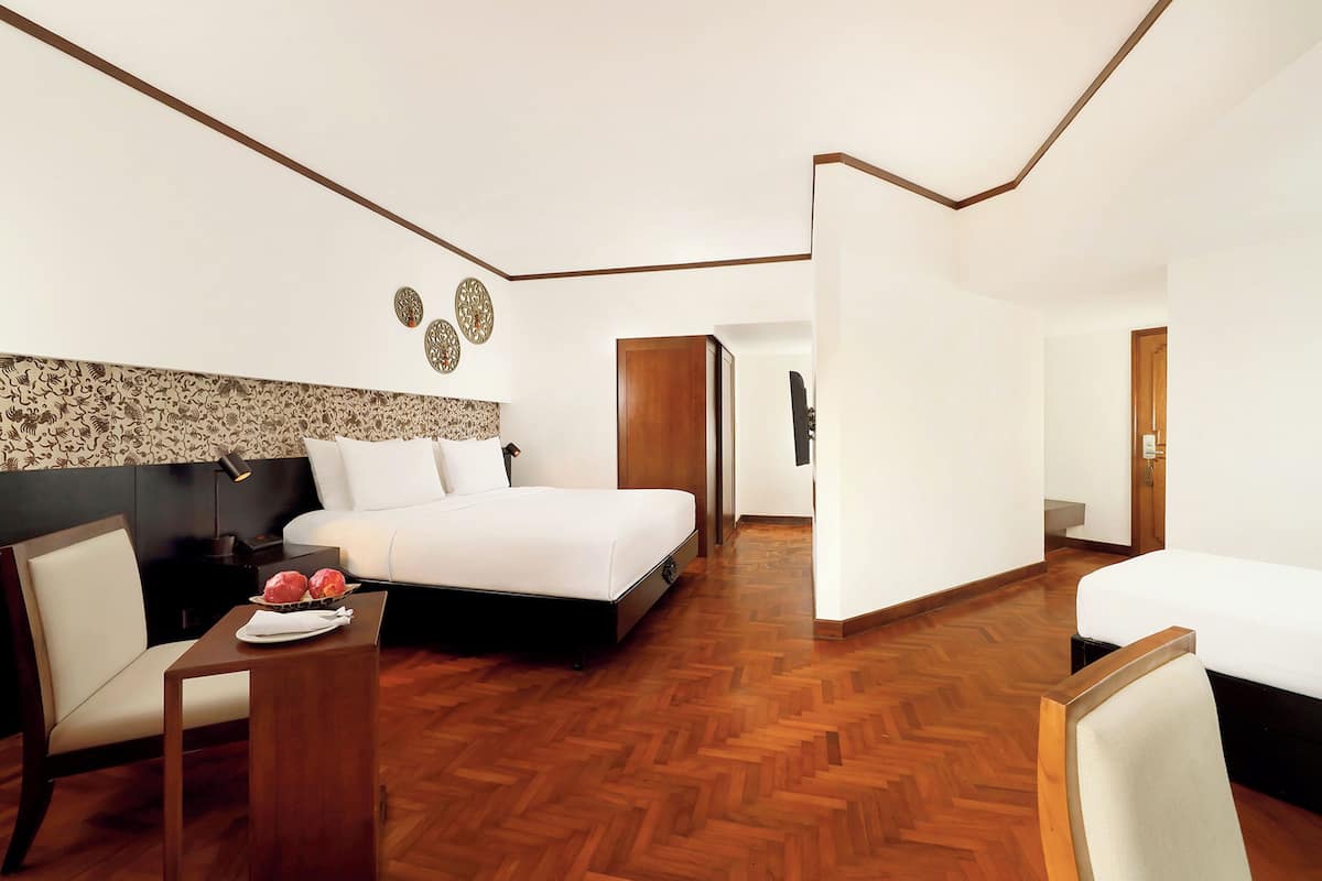 A modern hotel room with a large bed, a bedside table with a plate of fruit, a chair, and a wooden floor.