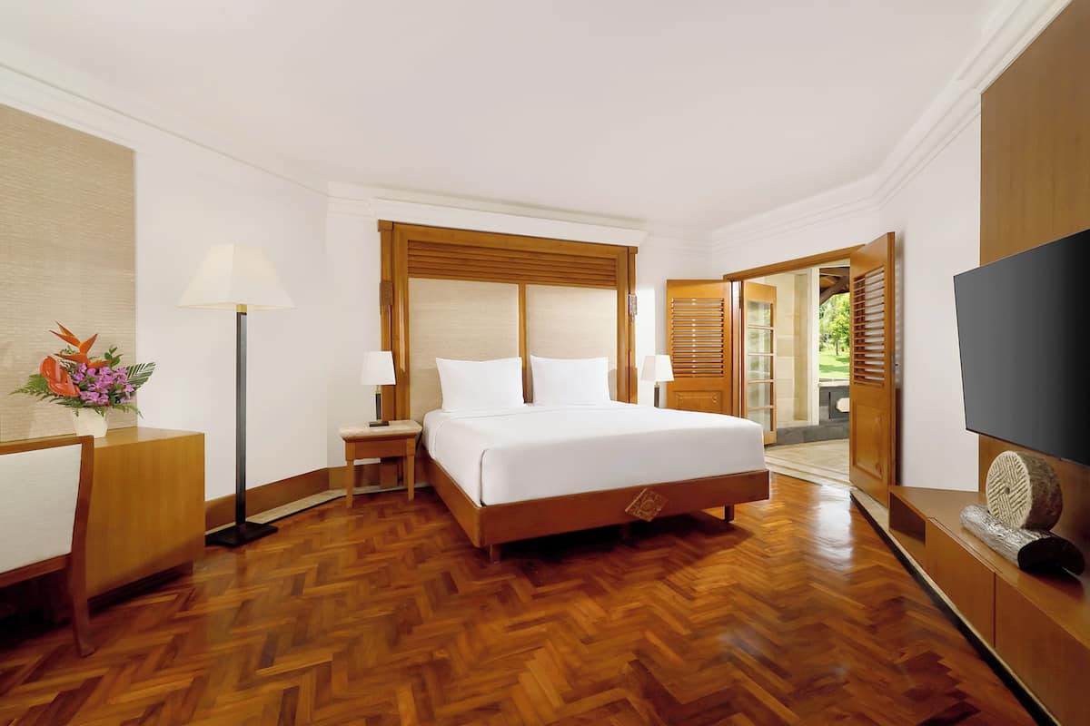 A hotel room with a large bed, wooden flooring, a desk with a lamp, a TV, and a view of the outdoors.