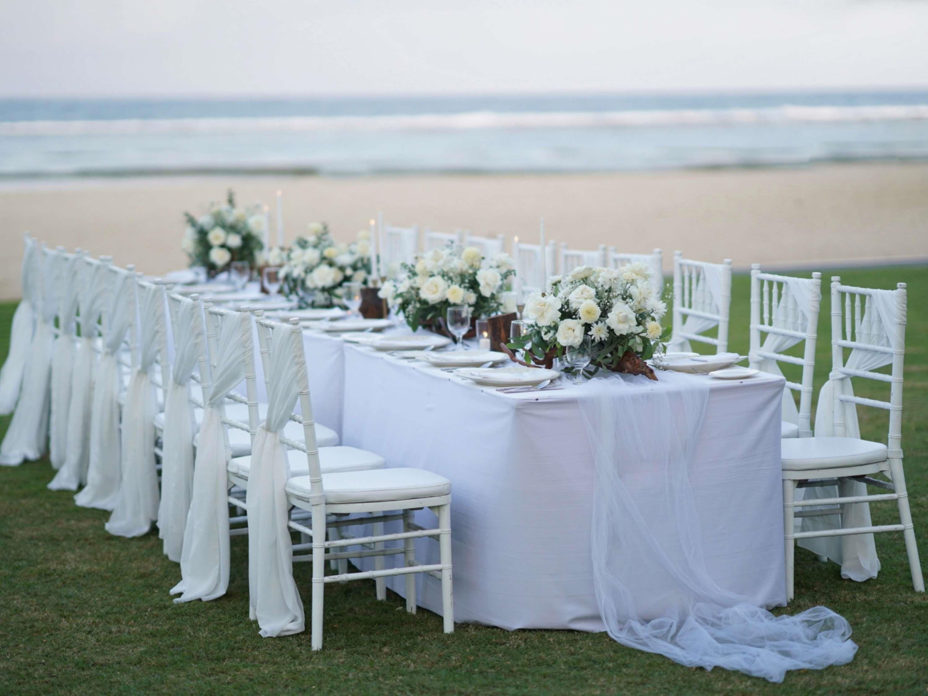 Una larga mesa dispuesta para una boda en la playa, con sillas blancas y centros de mesa florales.