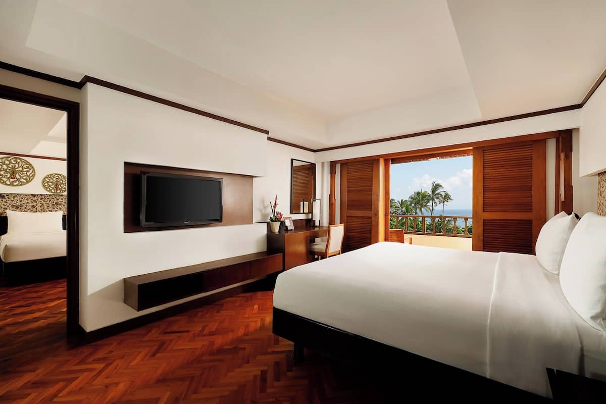 A modern hotel room with a large bed, a flat-screen TV, a desk, and a balcony with a view of palm trees.