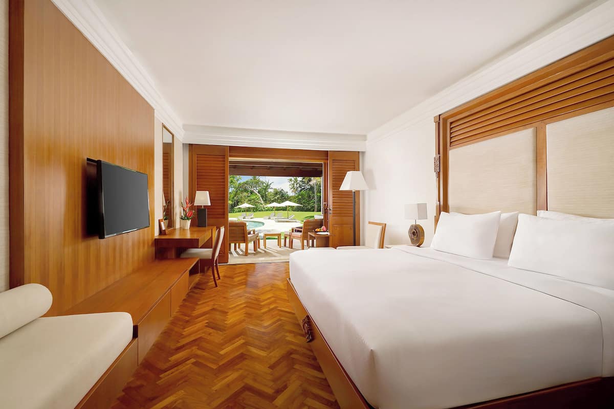 A hotel room with a large bed, a flat-screen TV, wooden paneling, a wooden floor, and a view of a garden and pool.