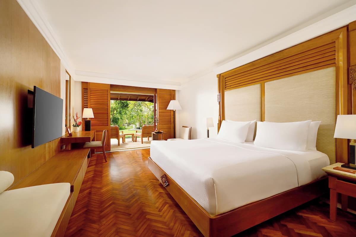 A hotel room with a large bed, wooden flooring, a television, a desk, and a view of greenery outside.