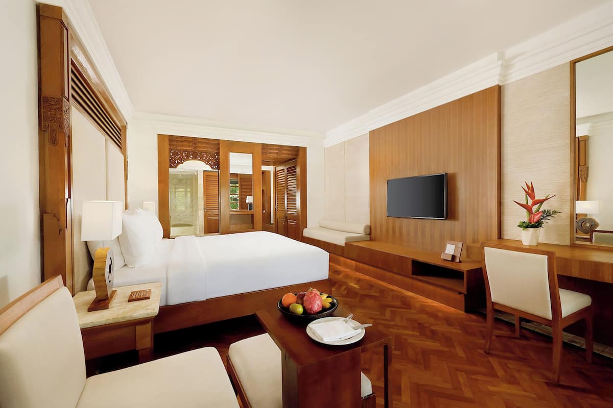A hotel room with a bed, a desk, a chair, a television, and a dining table with fruit.