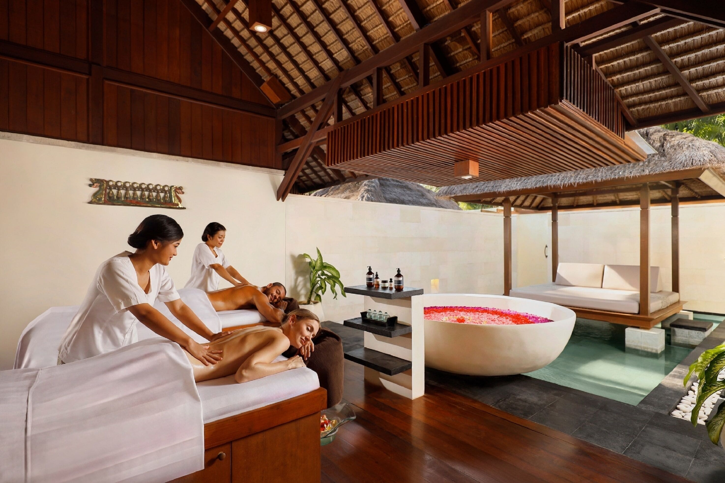 Uma área de spa com vigas de madeira, uma banheira grande com pétalas de rosa e uma mesa de massagem.
