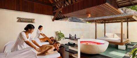 Uma área de spa com vigas de madeira, uma banheira grande com pétalas de rosa e uma mesa de massagem.