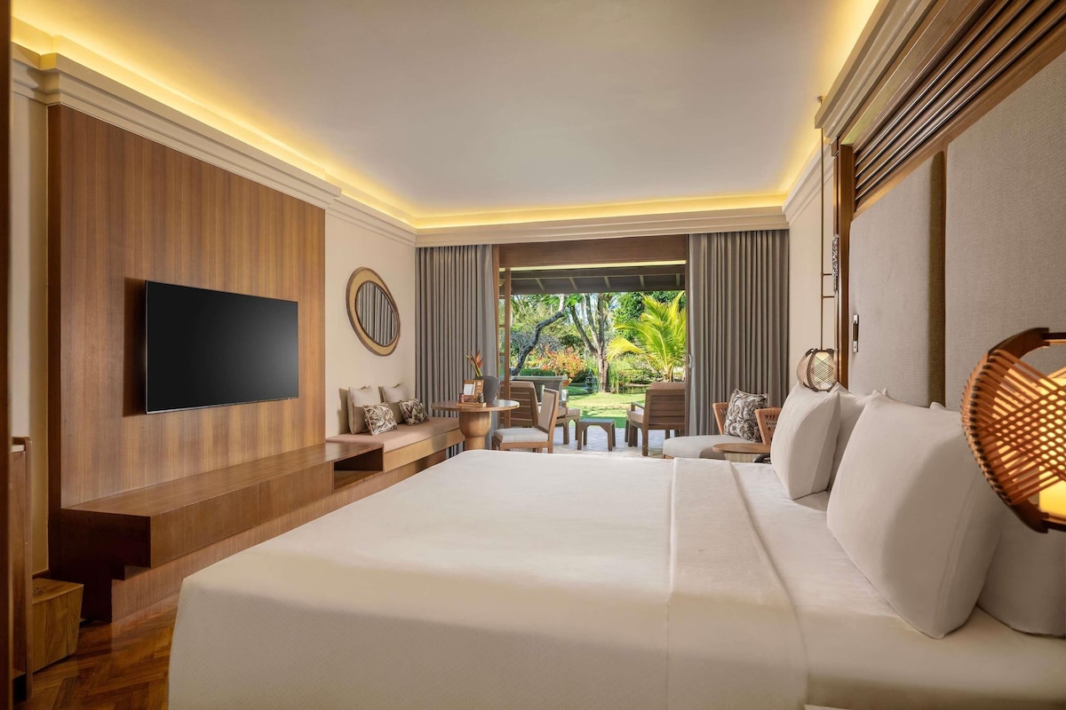 A hotel room with a large bed, a flat-screen TV, a seating area with a table and chairs, and a view of greenery outside.