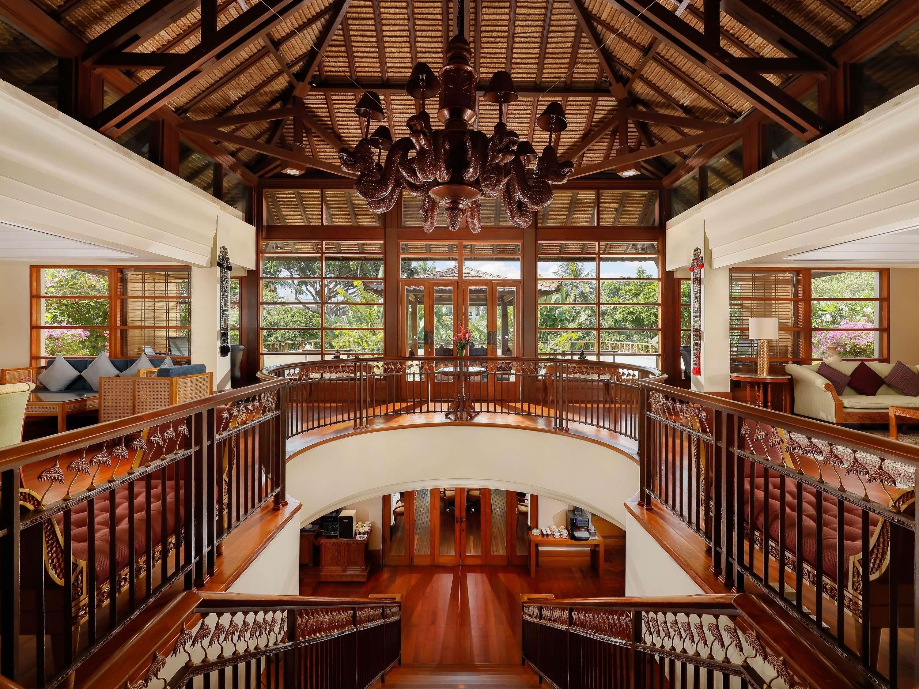 A spacious interior with a central staircase, wooden railings, and large windows offering a view of greenery outside.