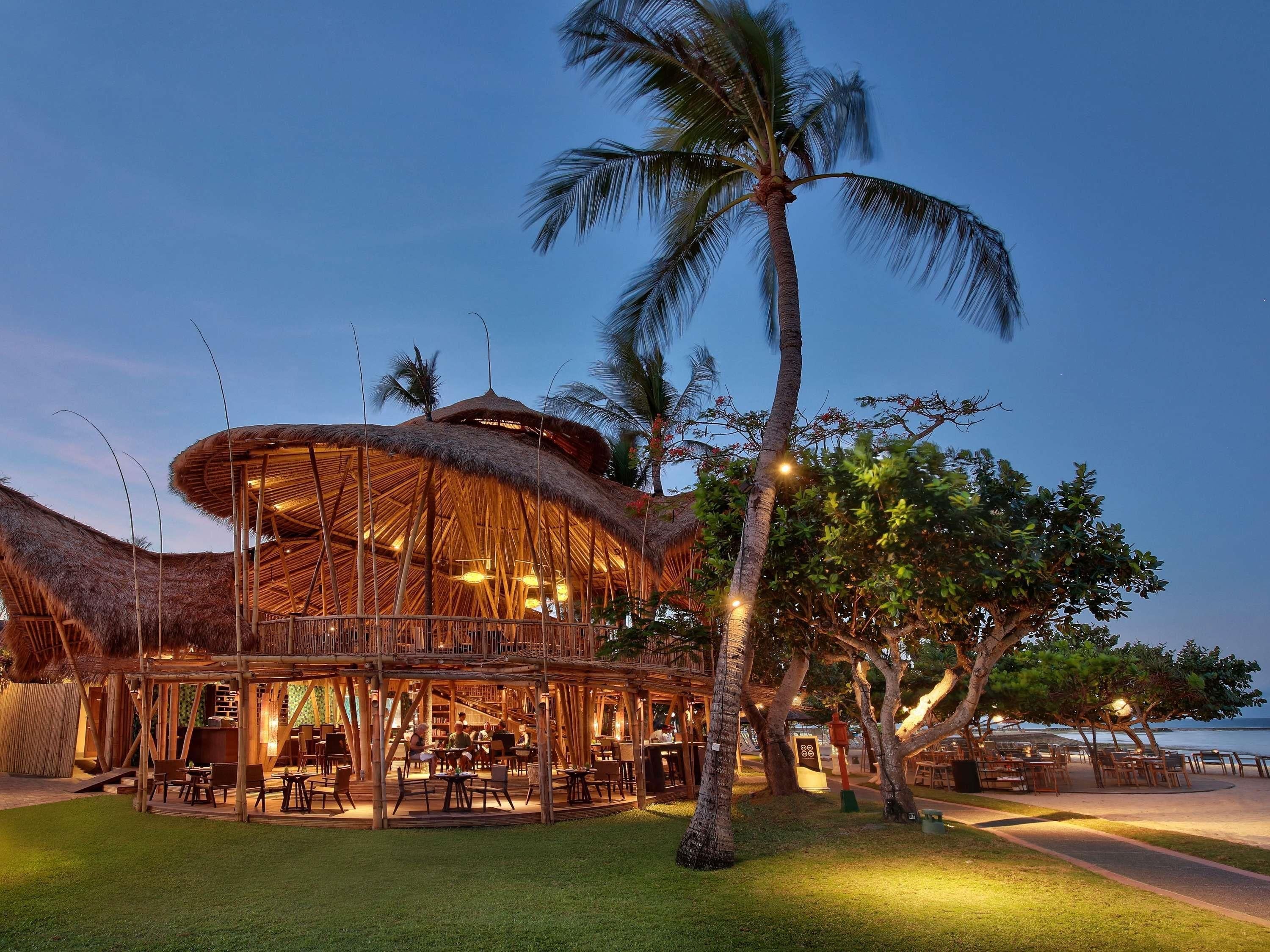 A thatched-roof outdoor dining area with tables and chairs, surrounded by palm trees and a view of the sea.