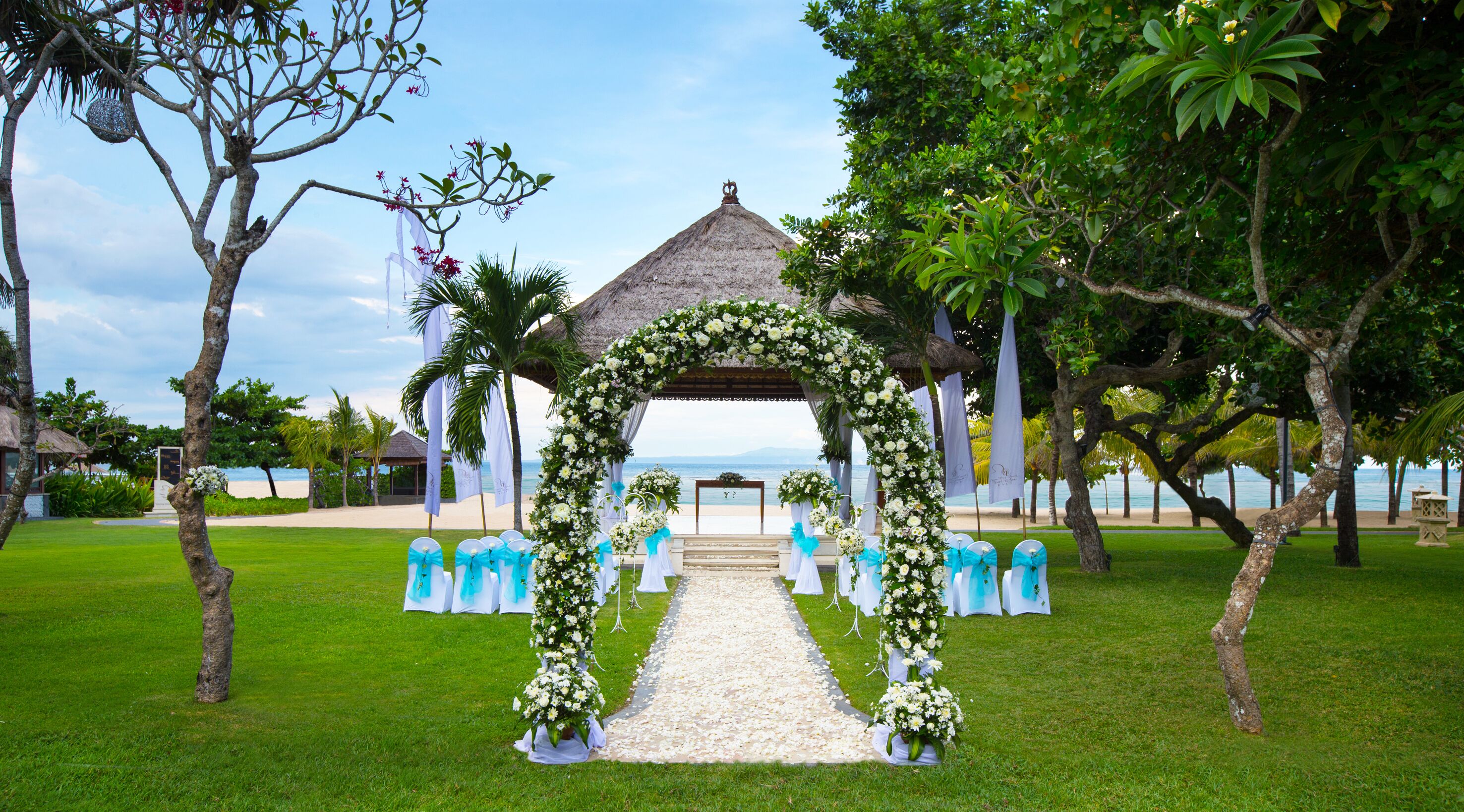 Une arche de cérémonie de mariage installée sur la plage, avec un toit de chaume, entourée de verdure et offrant une vue dégagée sur l’océan.