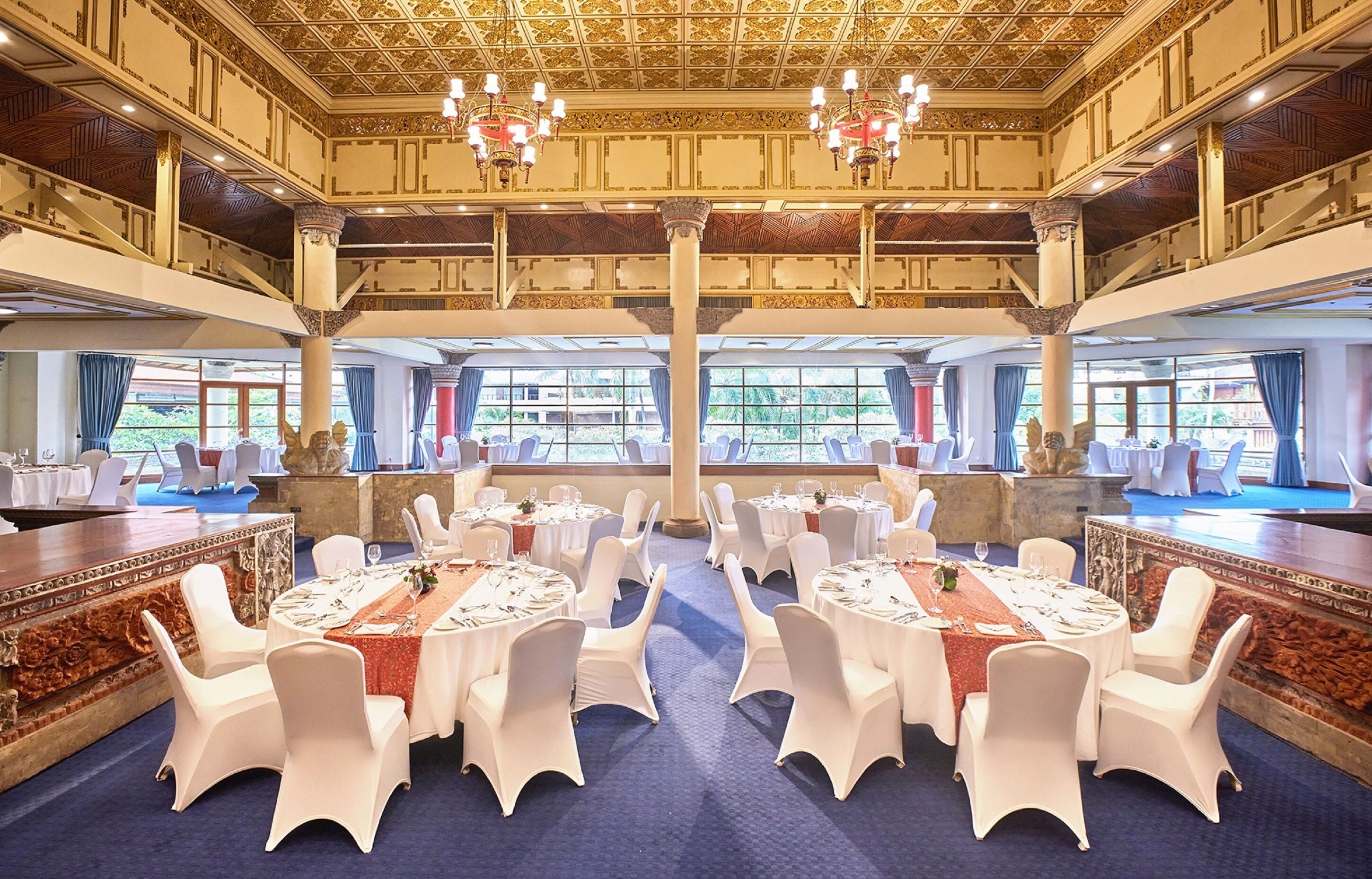A large banquet hall with multiple round tables set for dining, white chairs, and a grand chandelier.