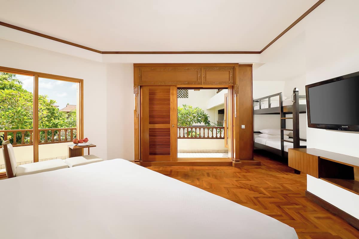 A modern hotel room with a large bed, a TV, a balcony with a wooden railing, and a view of greenery outside.