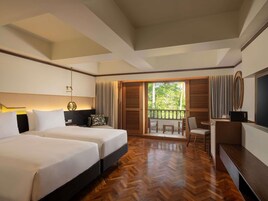 A hotel room with two beds, a desk, and a balcony with a view of trees.