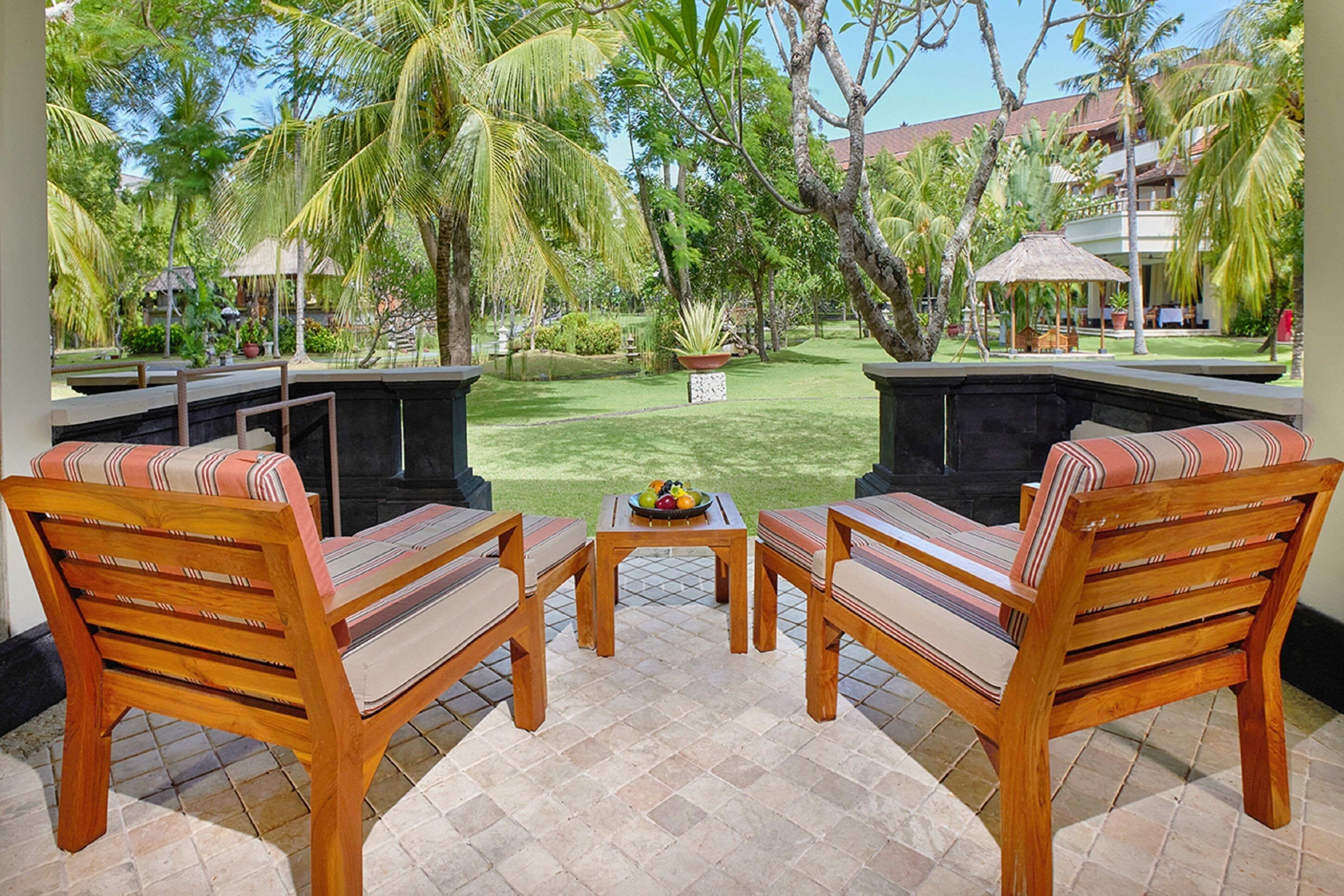 An outdoor seating area with wooden chairs and a table, surrounded by lush greenery and a view of a building in the background.