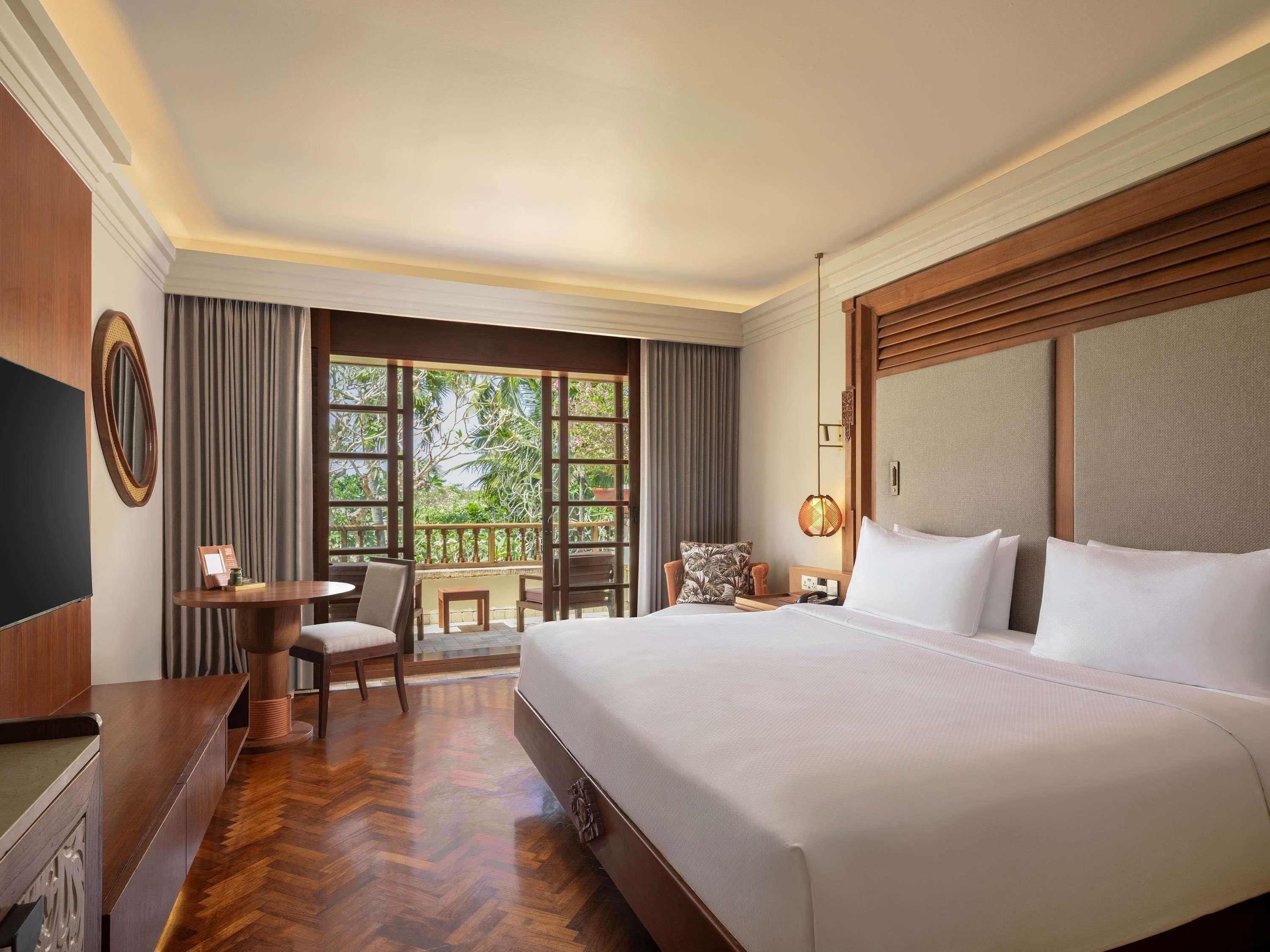 A hotel room with a large bed, a desk with a chair, a small table, and a view of greenery through the windows.