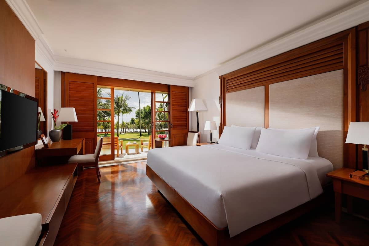 A spacious hotel room with a large bed, wooden furniture, and a view of palm trees.