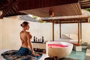 A person sitting on a mat in a room with a large white bowl filled with rose petals, a small table with bottles, and a white sofa. - Nusa Dua Beach Hotel and Spa - Handwritten Collection (Nusa Dua)
