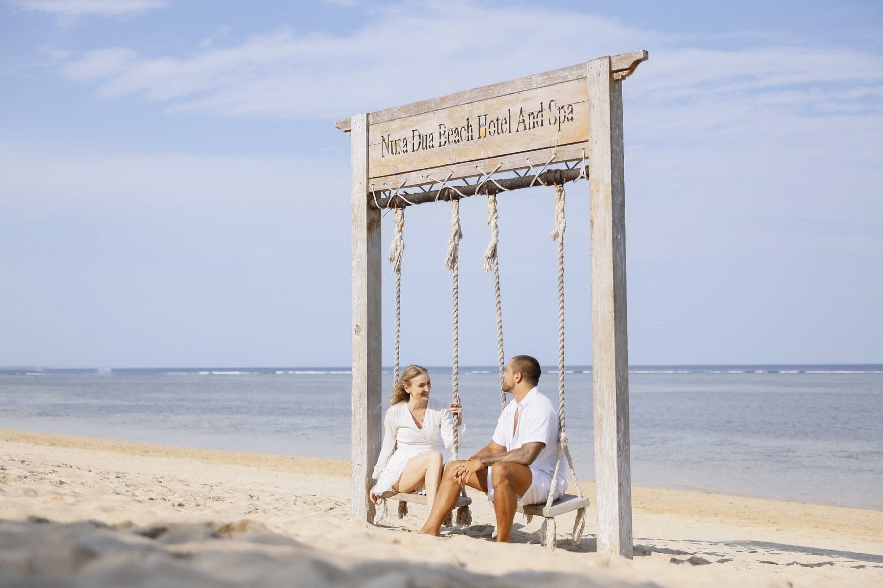 Een houten schommel met een bord waarop staat: "Nura Dua Beach Hotel And Spa" aan een strand.