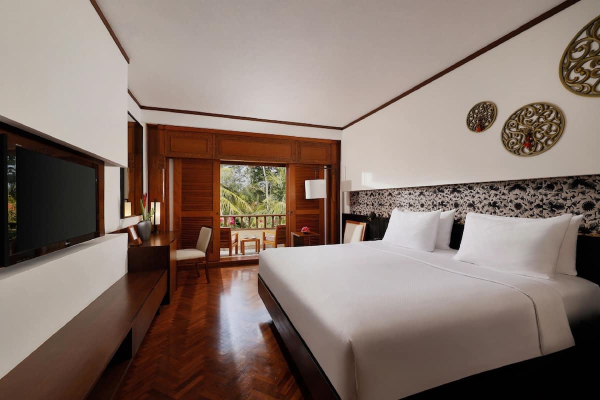 A hotel room with a large bed, a TV, a desk, and a balcony with a view of greenery.