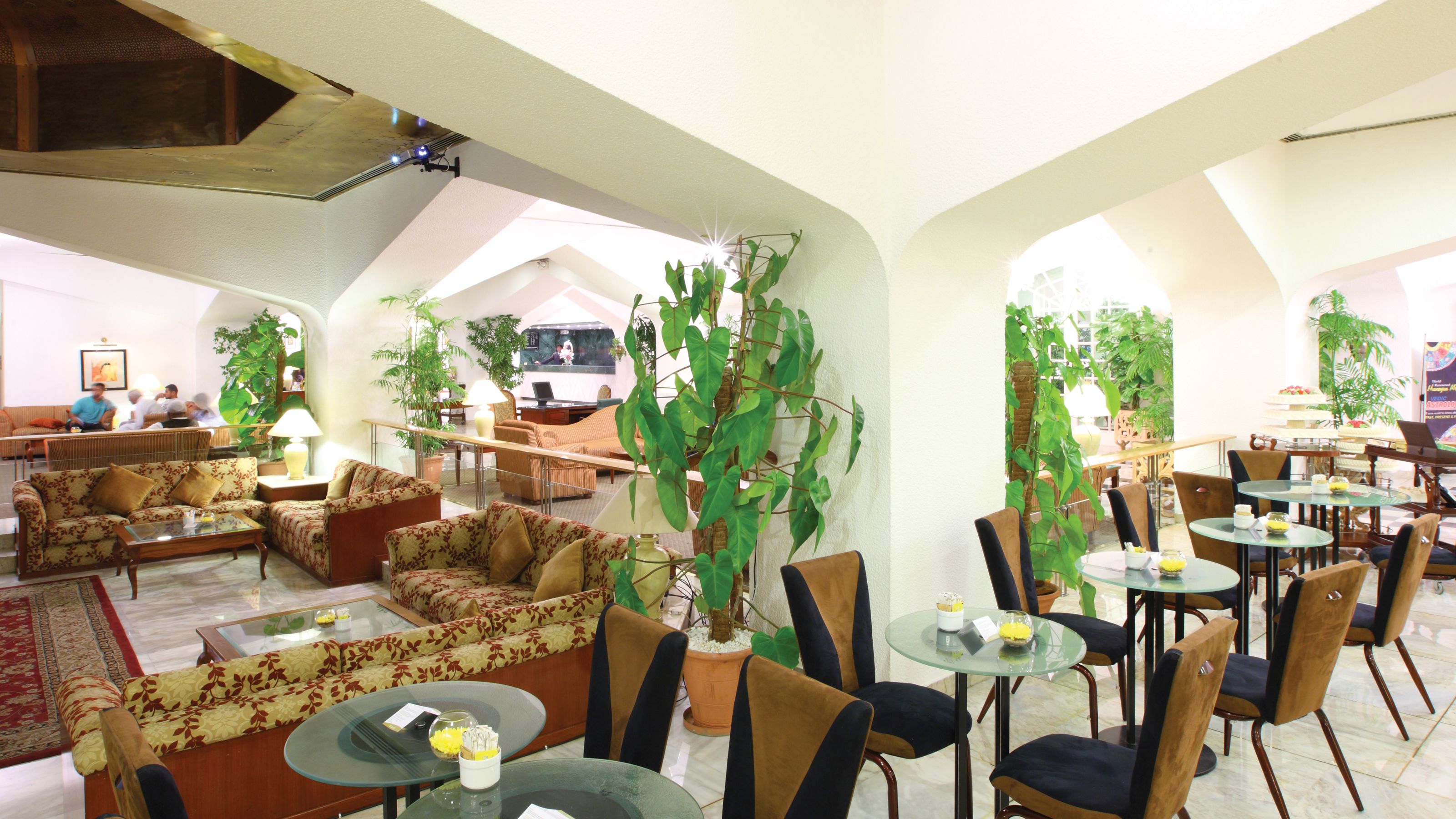 A spacious lobby with sofas, chairs, and tables, featuring large potted plants and a high ceiling with exposed beams.