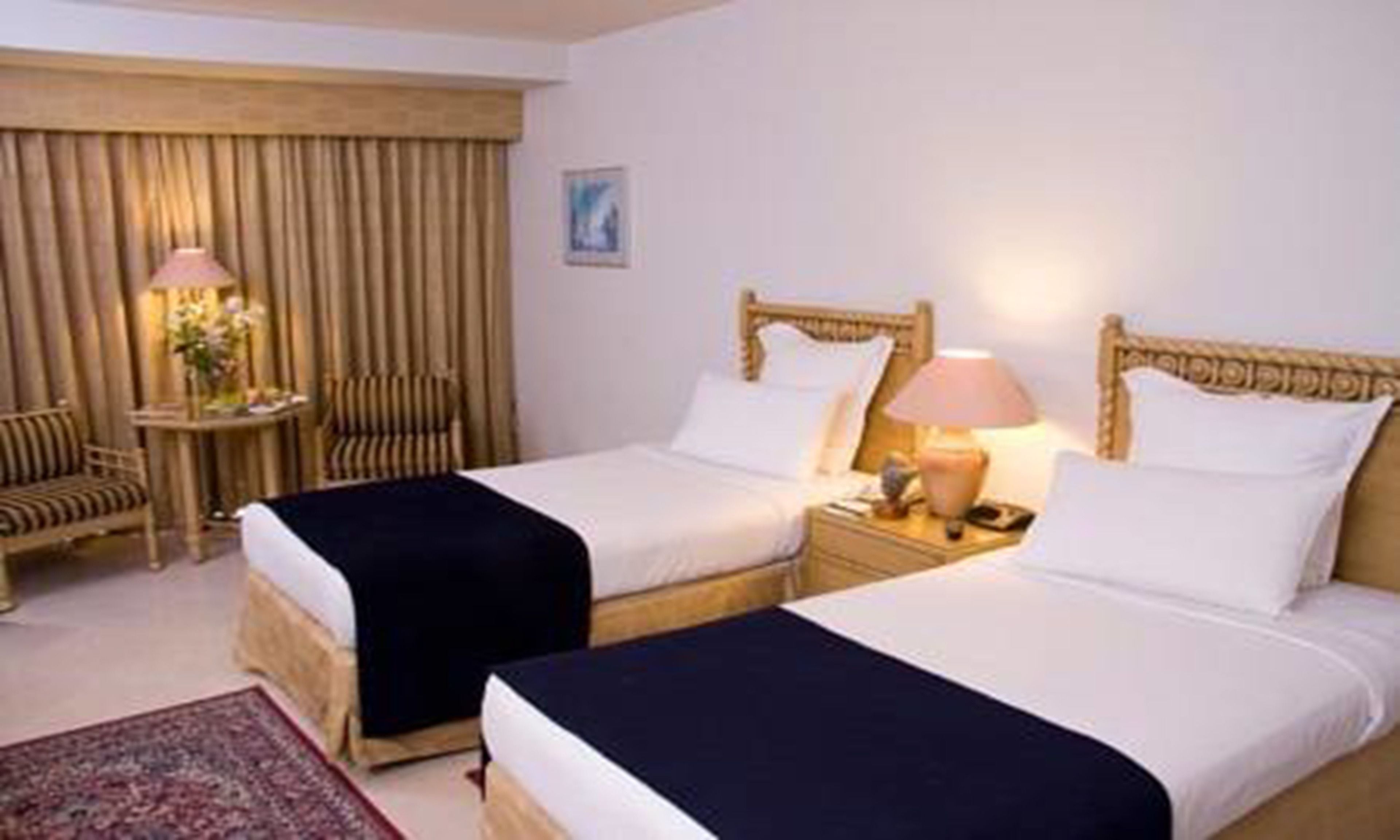 Una habitación de hotel con dos camas individuales, cabecero de madera, mesita de noche con lámpara, una silla y una pequeña mesa con un florero.