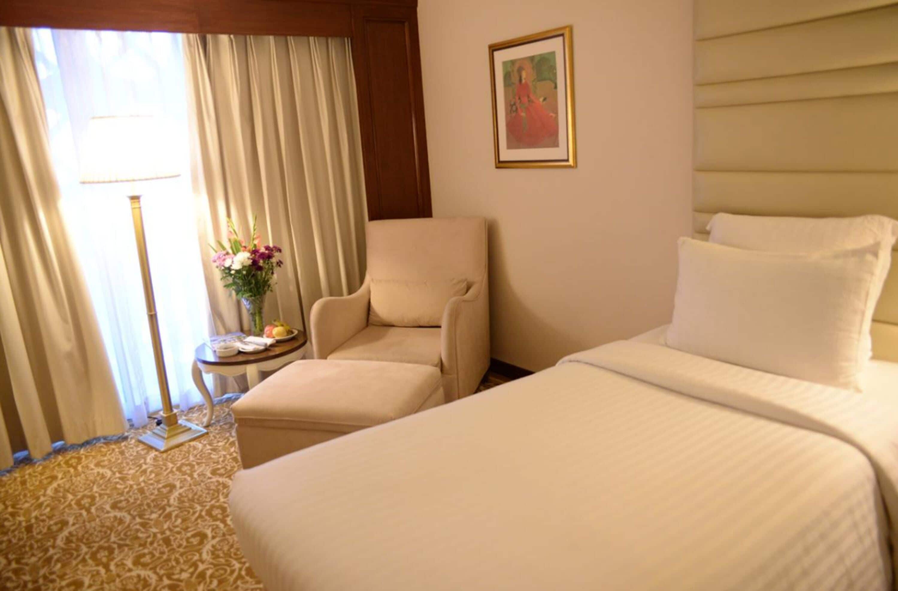 Una habitación de hotel con una cama, una silla, una mesita con un florero y un cuadro en la pared.